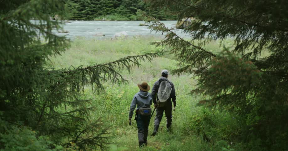 A view from behind shows a man and a woman in fishing gear walking away from the camera through a grassy clearing towards a wide river with pine trees lining the banks