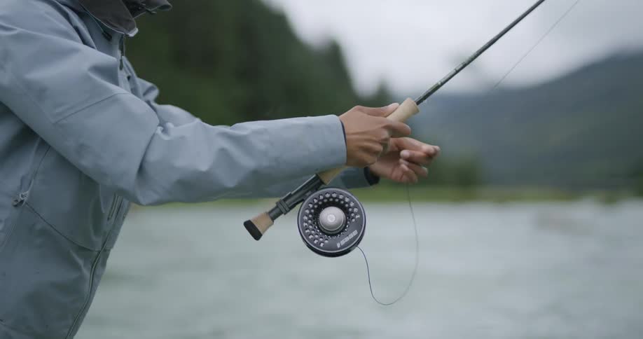 A close view of a persons hands holding a fly fishing rod and reel with a mountain and a river in the blurred background