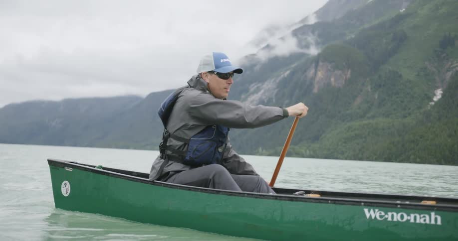 Man wearing a cap and life vest paddling a green canoe on a lake surrounded by mountains