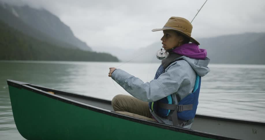 Woman in a green canoe fishing with a rod on a lake surrounded by mountains