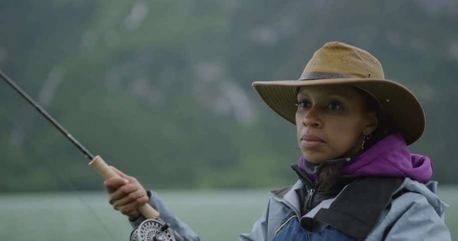 Woman in hat and raincoat fishing in a lake surrounded by mountains