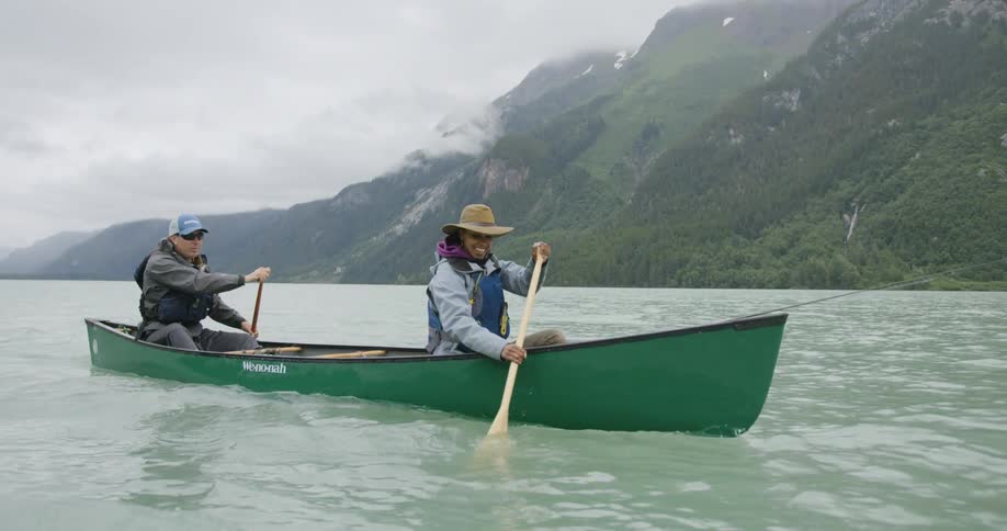 Two people paddle a green canoe on a lake surrounded by mountains