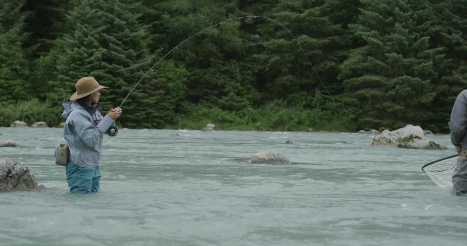 Woman fishing in a river with a bent rod and a partner holding a fishing net