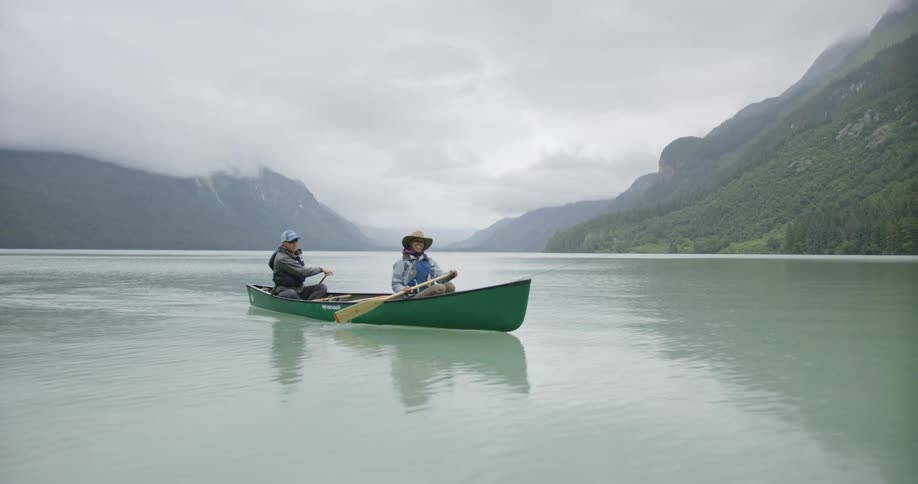 A man and a woman in outdoor gear are paddling a green canoe across a large calm lake surrounded by mountains under a cloudy sky