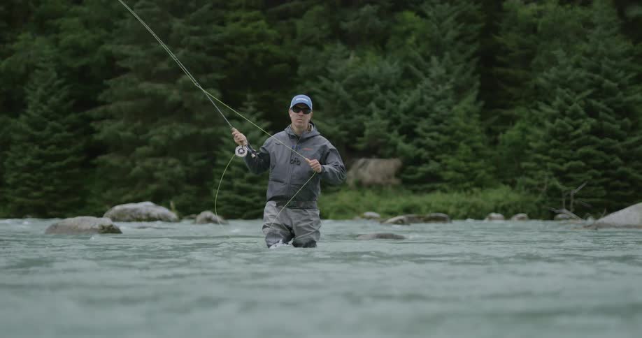 Fisherman with a blue cap and sunglasses fly fishing in a river surrounded by trees in the background