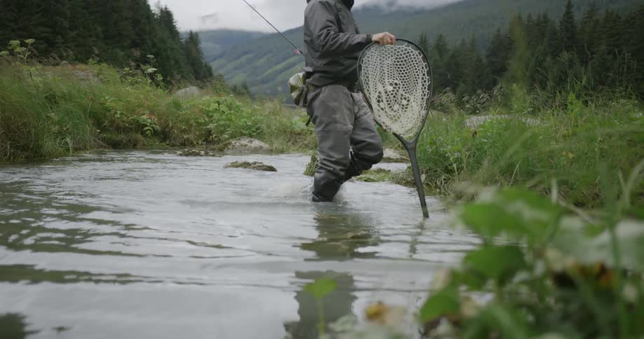 A person is walking through water holding a fishing net with a background of mountains and trees