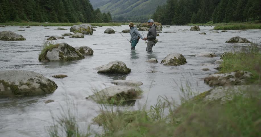 Two people fishing in a shallow rocky river surrounded by vegetation and mountains in the background
