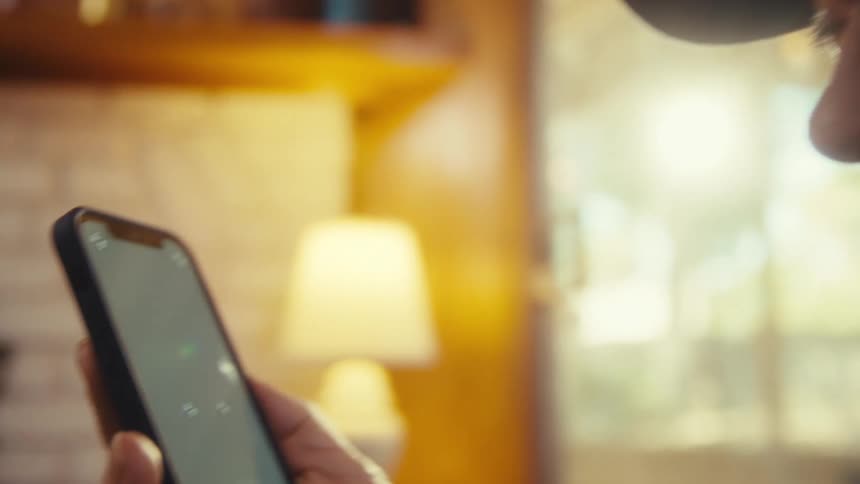 A closeup side profile shot of an older mans face is shown with a lamp glowing in the background his expression is serious as he looks at something the shot then cuts to a hand holding a modern smartphone