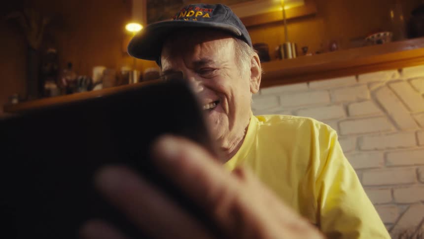 An older man wearing a worlds best grandpa hat and a yellow tshirt laughs as he looks at a tablet he is seated in a sunlit room in front of a fireplace with a clock on the mantle