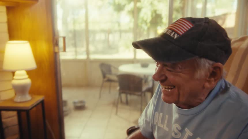 Elderly man wearing a blue shirt and a baseball cap looking at a smart phone and laughing while sitting down on an armchair indoors during day time