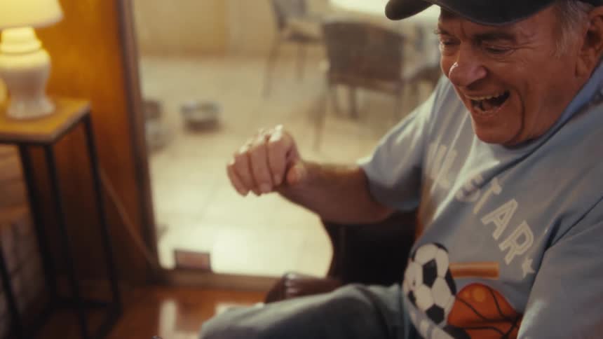 Elderly man wearing a baseball cap cheering while looking at the screen of a smart phone indoors during day time