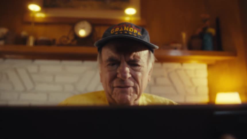 An elderly man is looking and reading at a black item he is holding in his hands while in a softly lit living room
