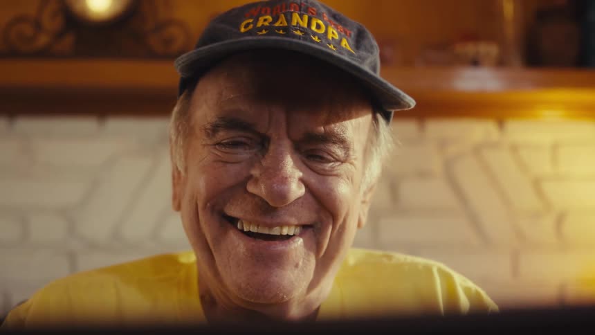 A shot from a low angle shows an elderly man wearing a black baseball cap that says worlds best grandpa he smiles widely as he looks down at a screen possibly a tablet or laptop