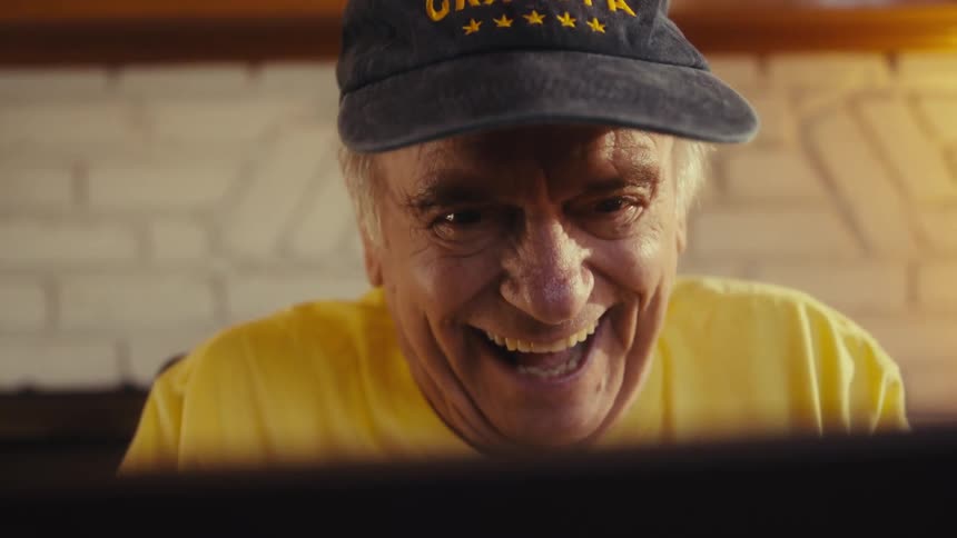 Elderly man wearing a yellow shirt and a baseball cap laughing in front of an electronic device indoors during day time