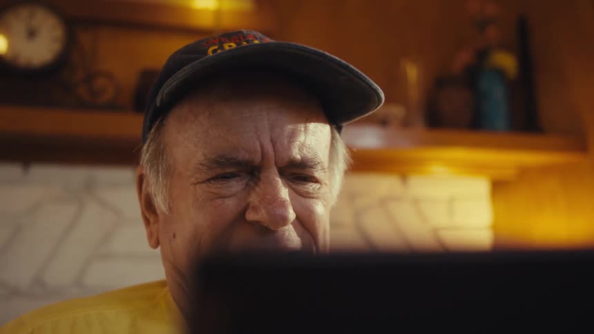 Elderly man wearing a baseball cap and a yellow shirt sitting in front of a laptop angry and nervous before laughing indoors during day time