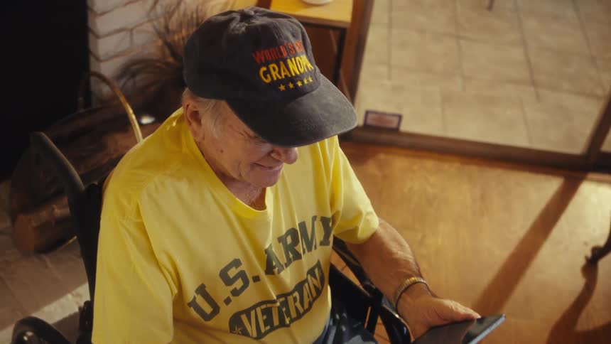 A closeup shot shows a man in a wheelchair looking down at a tablet hes wearing a yellow tshirt that says us army veteran and a baseball cap that reads worlds best grandpahe smiles warmly as he looks up from the tablet suggesting hes watch