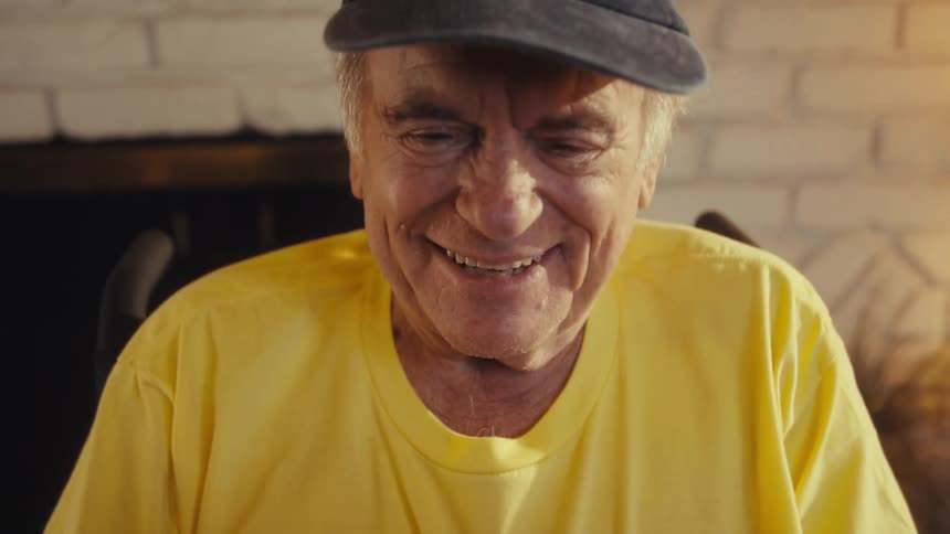 Elderly man sitting down wearing a yellow shirt and a baseball cap laughing while holding a tablet on his hands indoors during day time