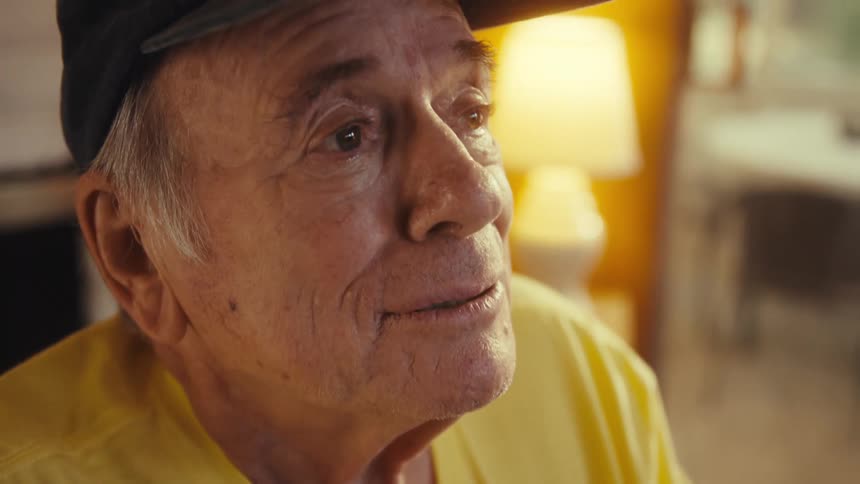Elderly man wearing a baseball cap and a yellow shirt sitting down and smiling in a living room indoors during day time