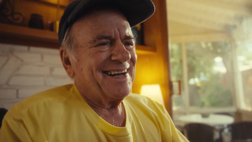 Elderly man sitting wearing a yellow shirt and a cap holds a tablet with both hands while looking at the screen with a gentle smile