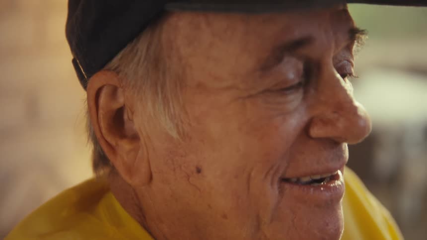 Elderly man wearing a baseball cap and a yellow shirt talking and laughing indoors during day time