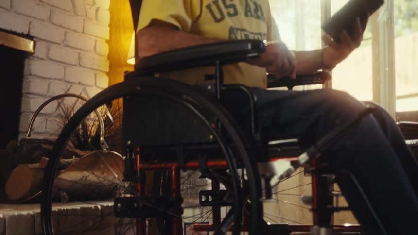 Elderly man in a wheelchair smiles broadly while holding a tablet illuminated by sunlight streaming through the window