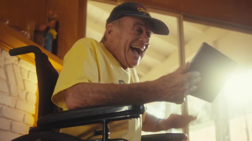 An elderly man sits in a wheelchair in a sunlit room holding a tablet he looks down at the screen then his face breaks into a wide smile as he begins laughing and raising his hand in excitement as if seeing a loved one