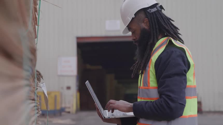 Warehouse worker standing near a tall stack of wood uses a laptop