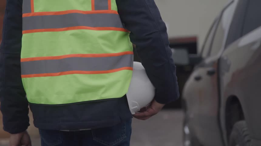 A worker in a safety vest walks with a hard hat beside a vehicle