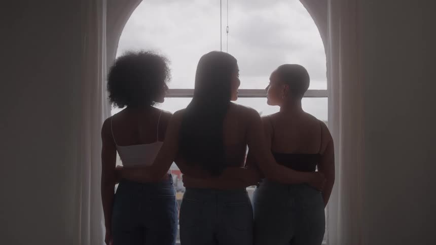 A rearfacing silhouette shot shows three women standing with their arms around each other looking out a large window their bond is emphasized as they stand together united in a shared moment