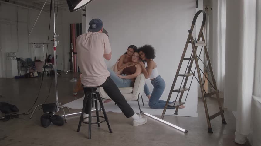 A wide shot of a brightly lit professional photo studio shows a male photographer sitting on a stool hes taking a picture of three female models one of whom is sitting in a chair and the other two kneeling beside her
