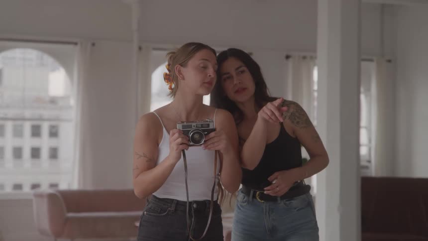 A woman holds a camera and points it to the front while another woman stands beside her and points at the camera