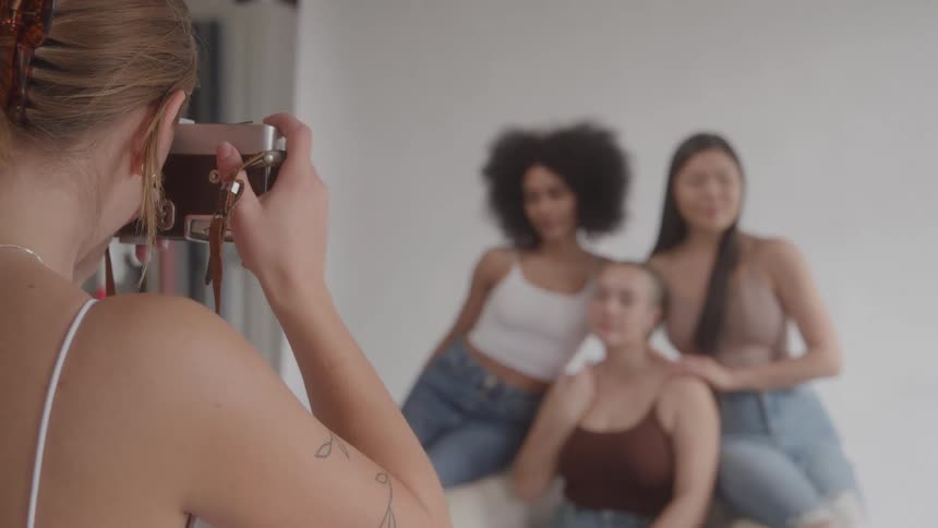 A female photographer is shown from a rear angle holding a vintage camera to her eye the shot is in focus on the photographer while the three women she is taking a picture of are blurred in the background
