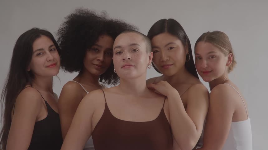 A group of five ethnically diverse women including one with a shaved head and another with an afro pose closely together against a plain studio background looking at the camera with gentle confident smiles