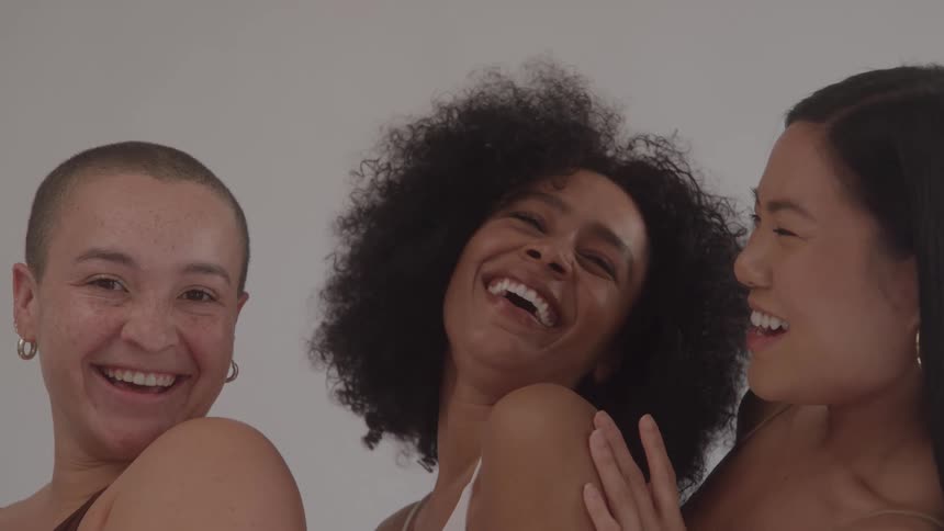 Three ethnically diverse womenone with a shaved head one with an afro and one east asianshare a moment of genuine joy laughing and smiling together during a candid studio photoshoot against a plain background