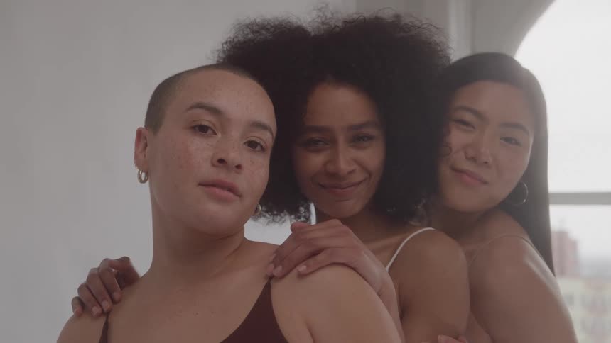 Three young women smile as they pose together near a large window