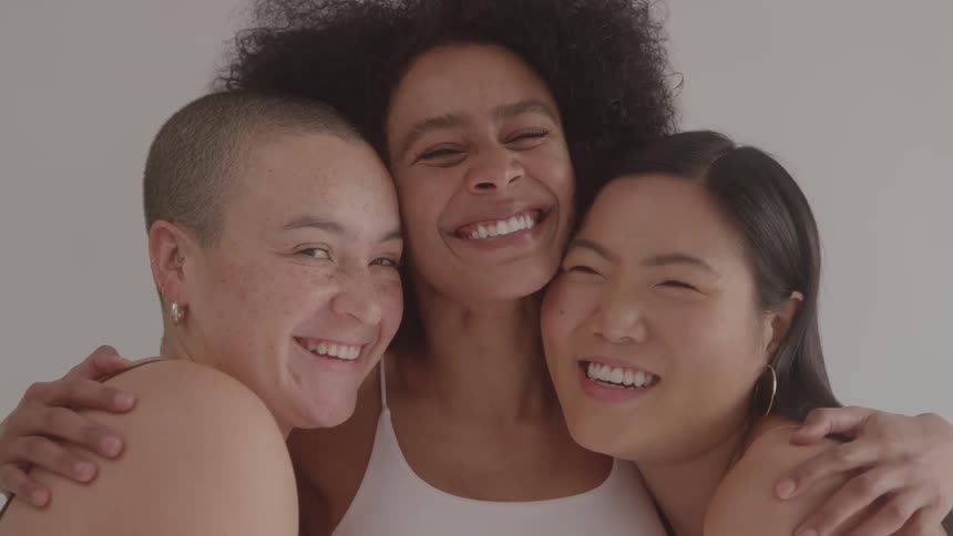 A group of three diverse women with a black woman in the middle smile and laugh together as they wrap their arms around each other the women are enjoying each others company as they all look into the camera with big smiles