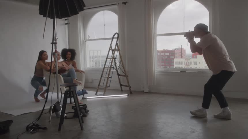 This behindthescenes video shows a male photographer actively shooting three diverse female models as they pose dynamically around a chair in a professional sunlit studio with a large white backdrop