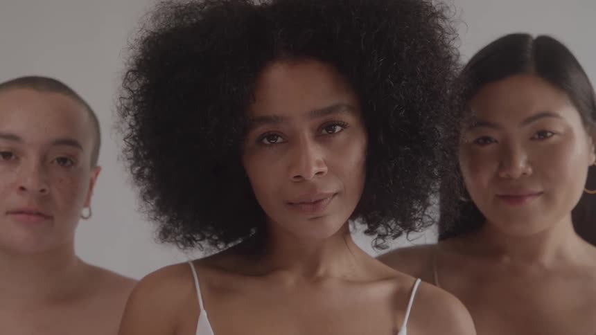 This is a video of three women with diverse features standing in a studio the camera focuses on the woman in the center who has curly hair and she closes her eyes before looking directly at the camera the other two women are out of focus on either sid