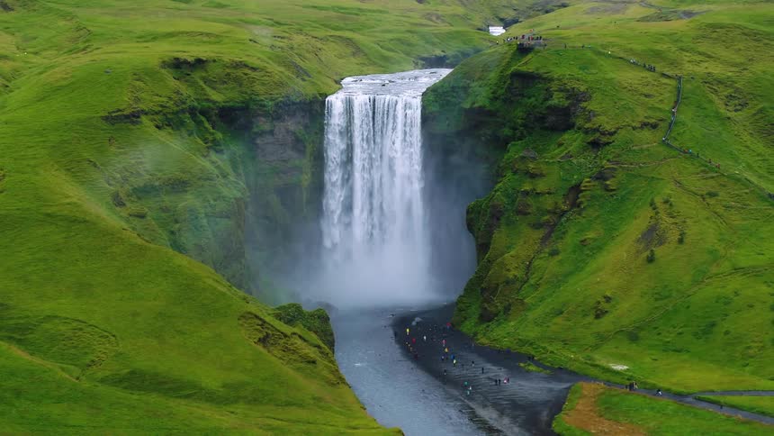 A breathtaking wide shot shows a stunning waterfall cascading down a cliff between two large lush green hills a group of tourists are visible at the bottom of the falls appearing tiny in comparison to the massive landscape