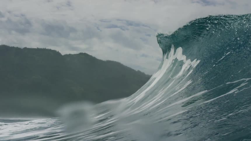 A shot from the waters perspective shows a large powerful ocean wave cresting and beginning to crash down the camera dips underwater as the wave passes over it and we see a small dark shape possibly a fish swimming away