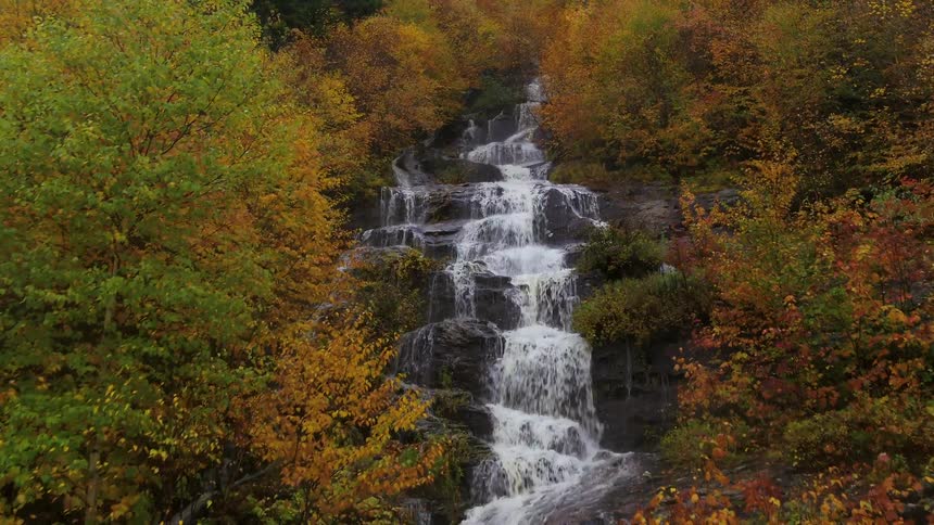 A stunning waterfall flows over a rocky cliffside surrounded by trees and bushes with beautiful fall foliage the shot is taken from a drone showing the entire height of the falls and the forest on either side of the river below