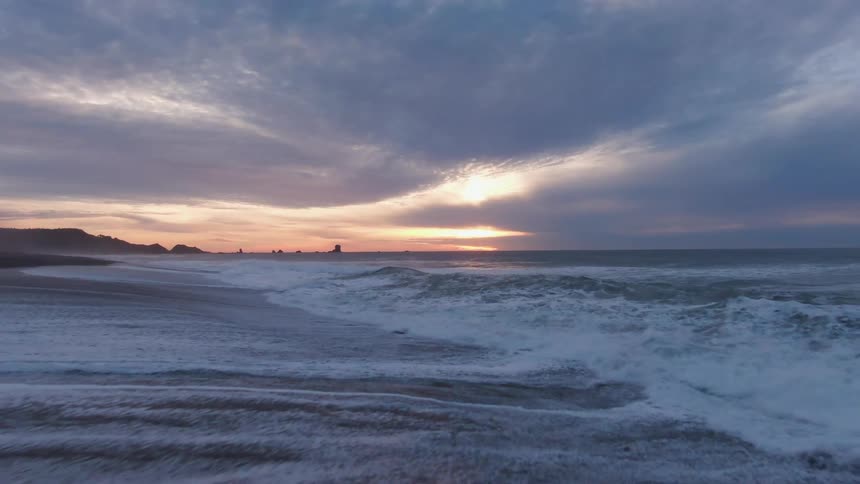 The video shows a powerful ocean with whitecapped waves crashing against a dark sandy beach at sunset the sky is filled with dramatic clouds with the suns golden light peeking through to create a bright reflection on the water