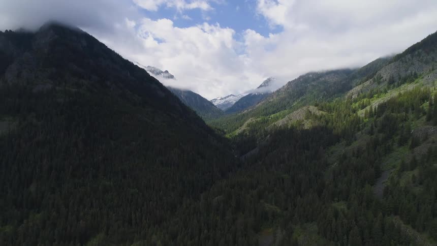 A majestic aerial shot slowly pulls backward out of a deep mountain valley revealing the vast landscape of dense evergreen forests a winding river below and distant snowcapped peaks partially covered by clouds