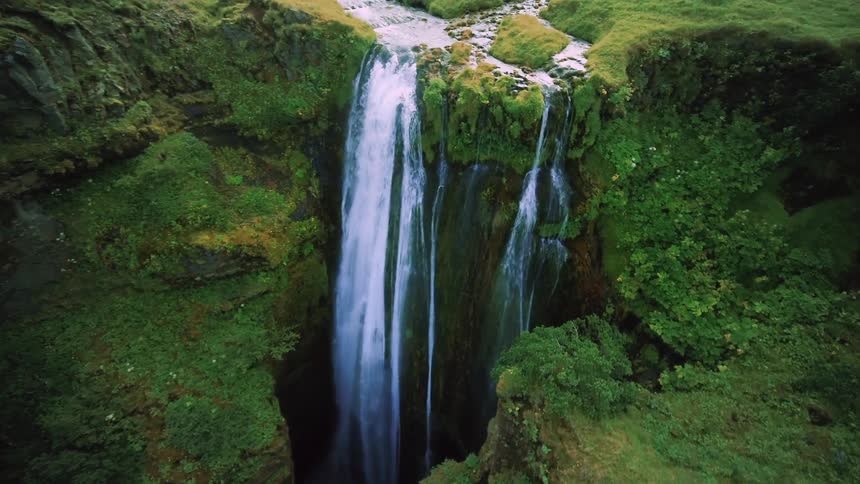 A tall narrow waterfall cascades down a rocky cliff face covered in lush green vegetation the water splits into multiple streams as it falls surrounded by moss and small plants that thrive on the misty rock