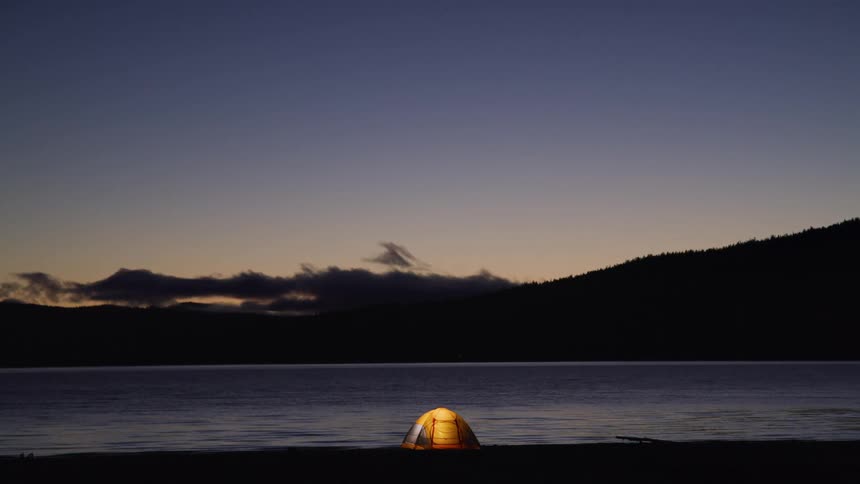 In a serene static shot at twilight a single small tent glows with a warm yellow light from within pitched in peaceful solitude on the shore of a vast calm lake surrounded by dark silhouetted mountains
