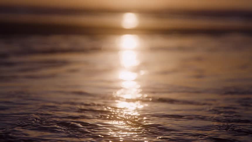 A mesmerizing closeup view lingers on the waters surface as the golden light from a setting sun creates a brilliant shimmering reflection that is gently distorted by the continuous soft ripples of the waves