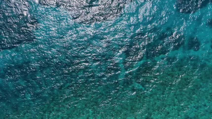 The video shows a highangle shot of crystalclear ocean water with a beautiful gradient from deep blue to bright turquoise the sunlight glitters on the surface as small waves and ripples move over the shallow rocky seabed038