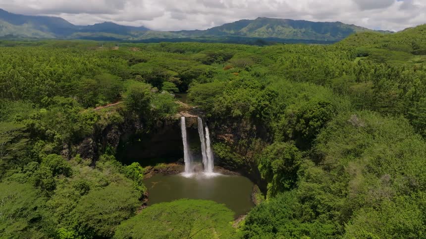 A stunning shot shows a massive twin waterfall cascading into a pristine green pool at the bottom of a cliff the surrounding landscape is a vibrant green forest with rolling hills and mountains visible in the distance under a bright sky050