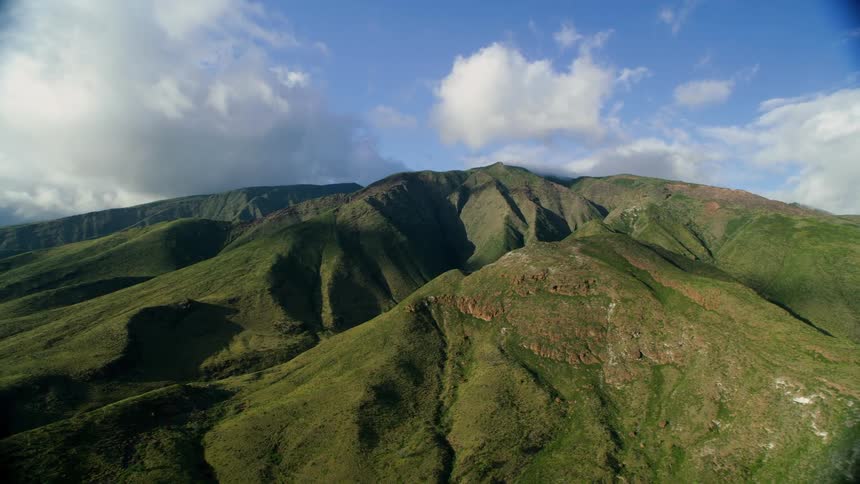 An aerial view shows a stunning mountain range covered in lush green foliage under a blue sky with scattered clouds the camera slowly moves revealing the detailed textures and deep valleys of the rugged landscape