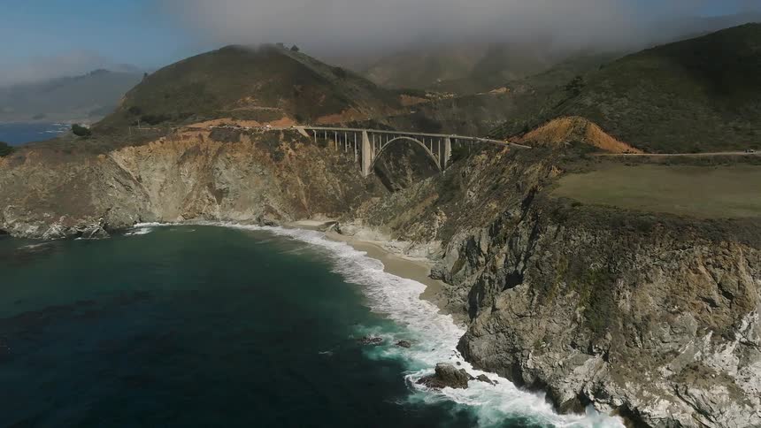 A shot shows a view of a bridge over a coastal landscape with mountains ocean and a beach